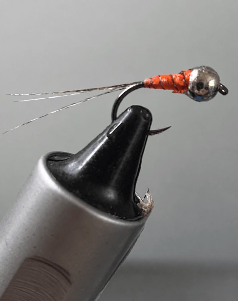 Close-up of a small fishing fly with a metallic bead head, orange body, and delicate hackle fibers, resting on a black pen.