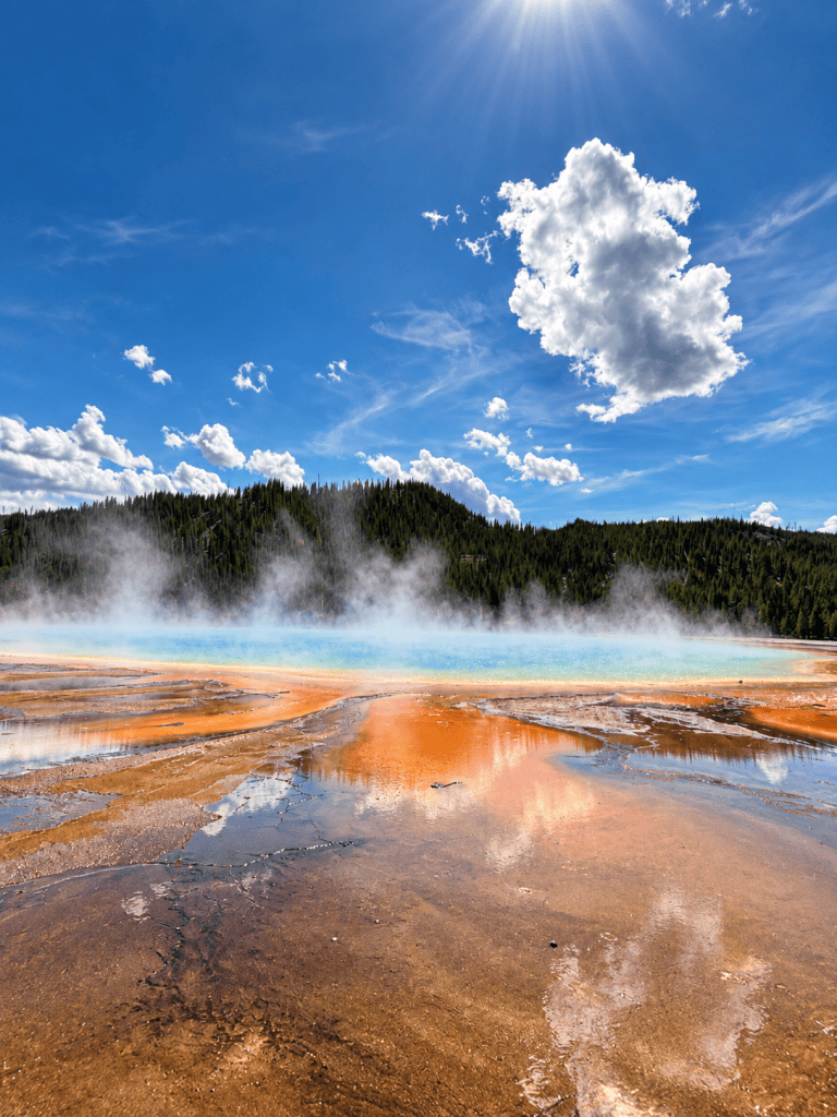 A vibrant geothermal spring surrounded by colorful mineral deposits and steam, with a bright blue sky and fluffy white clouds above.